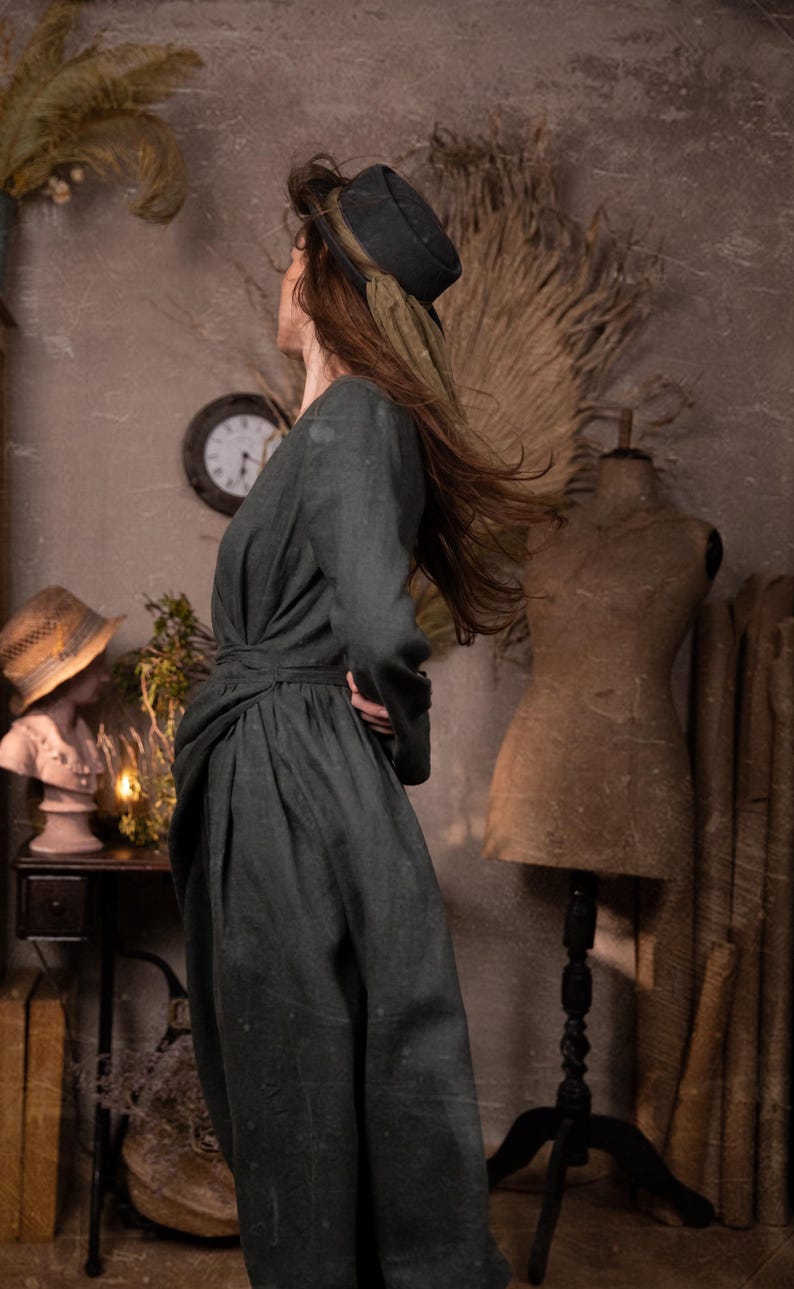 A person in a dark green outfit and a black hat with a green ribbon stands in a vintage-style room, facing away. The setting includes a mannequin, a straw-hat-wearing bust, a clock, and dried plants.