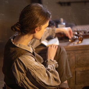 SPARROW linen vest in brown, shown on a seated model in a back three-quarter mid-length photo, with a sleeveless cut, plain back panel, wide arm opening, layered over a light gray blouse with gathered sleeves, Knock Knock Linen.