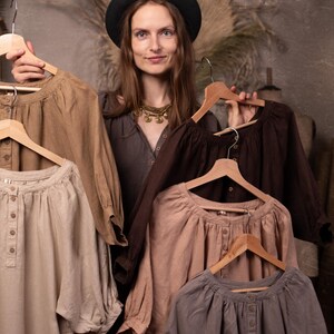 Linen dresses in neutral brown, beige, dusty pink, gray, and dark chocolate colors held on hangers by a model, front waist-up view, showing gathered necklines, front buttons, full sleeves, and soft folds.