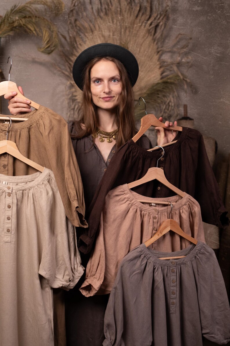 Linen dresses in neutral brown, beige, dusty pink, gray, and dark chocolate colors held on hangers by a model, front waist-up view, showing gathered necklines, front buttons, full sleeves, and soft folds.