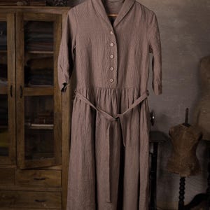 Linen dress displayed on hanger from a branch, front view full-length, folded collar with V opening, row of buttons, three-quarter sleeves, gathered skirt panel at waist, belt tied loosely with ends hanging; grey wall studio setting.