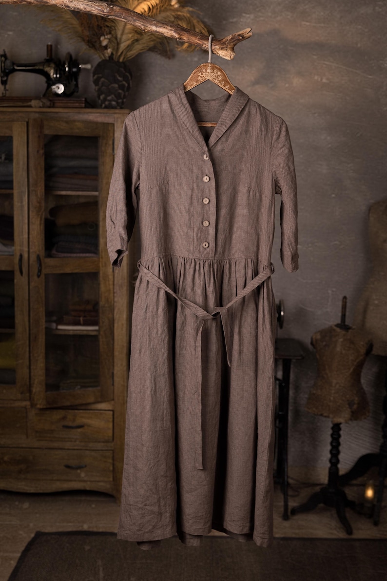 Linen dress displayed on hanger from a branch, front view full-length, folded collar with V opening, row of buttons, three-quarter sleeves, gathered skirt panel at waist, belt tied loosely with ends hanging; grey wall studio setting.