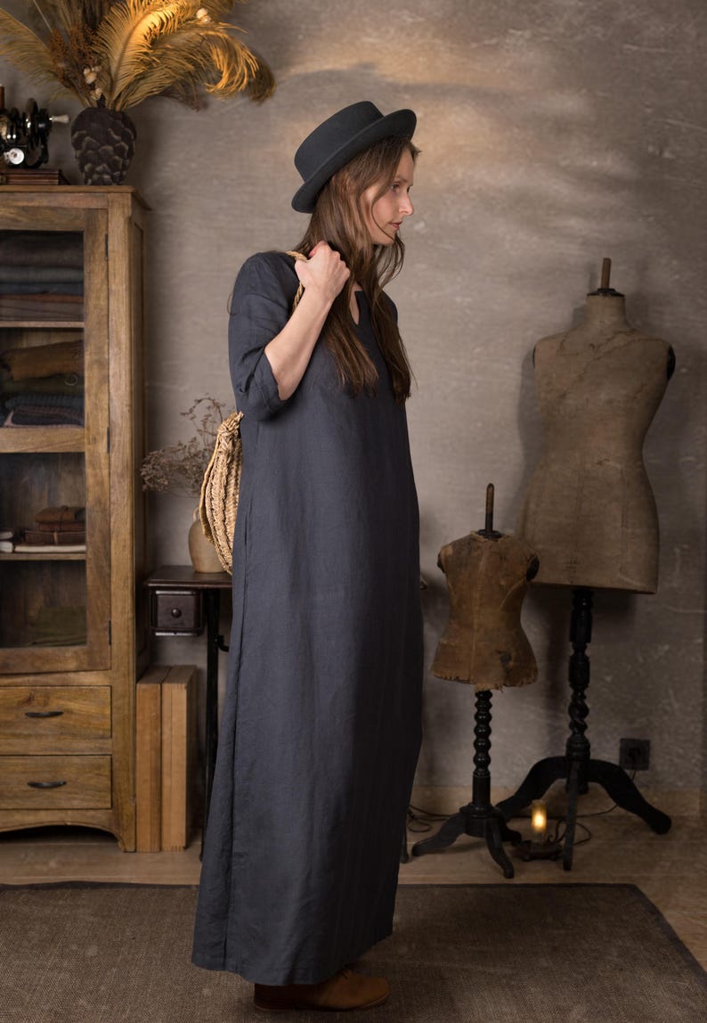 Woman in a long dark blue linen summer dress with 3/4 sleeves, shown from the side, wearing a black hat and holding a straw bag strap on her shoulder in a vintage-style room.
