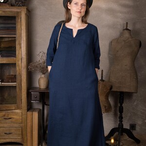 Model in a dark blue linen summer dress, ankle length with 3/4 sleeves and pockets, standing indoors against a neutral textured wall with wooden cabinet and dress forms.