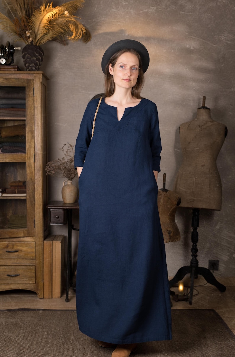 Model in a dark blue linen summer dress, ankle length with 3/4 sleeves and pockets, standing indoors against a neutral textured wall with wooden cabinet and dress forms.
