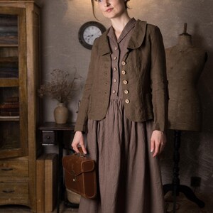 Woman in rustic studio wearing mushroom brown slim fit linen jacket with curved collar, long sleeves and button front over brown midi dress, holding brown leather briefcase, retro linen jacket by knock knock linen.