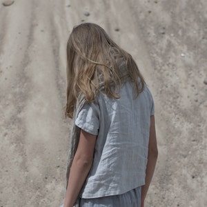 Back view of person standing on sandy ground, wearing a dusty sky short-sleeved buttoned linen top and matching loose pants. Hair is long and loose. Scene photographed outdoors with scattered rocks on sand. Linen blouse, buttoned shirt, loose linen top.
