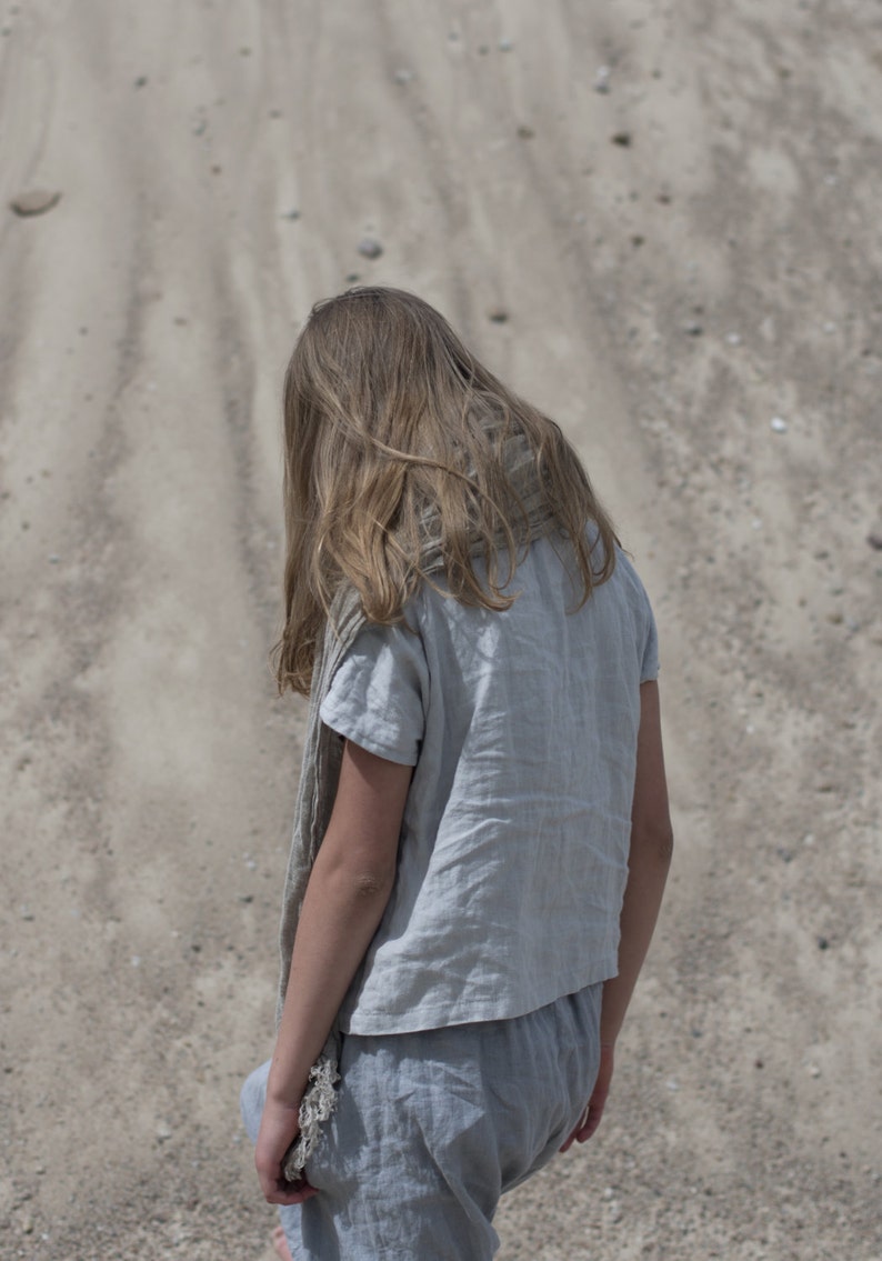 Back view of person standing on sandy ground, wearing a dusty sky short-sleeved buttoned linen top and matching loose pants. Hair is long and loose. Scene photographed outdoors with scattered rocks on sand. Linen blouse, buttoned shirt, loose linen top.
