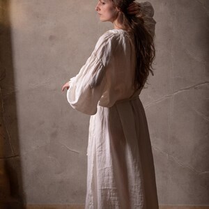 May include: A long-sleeved, floor-length white dress with a gathered neckline and voluminous sleeves. The garment features a simple, loose-fitting silhouette with a tie at the waist, crafted from a textured, opaque fabric that falls to the ankles.