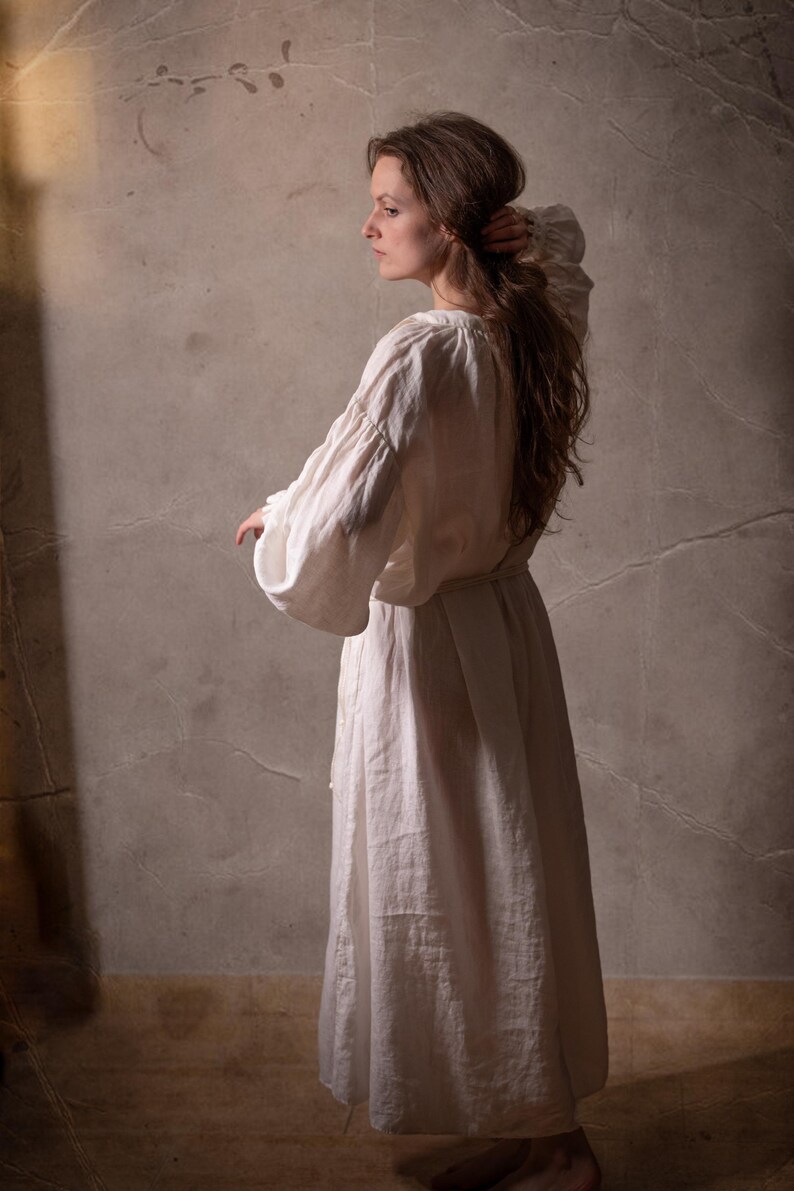 May include: A long-sleeved, floor-length white dress with a gathered neckline and voluminous sleeves. The garment features a simple, loose-fitting silhouette with a tie at the waist, crafted from a textured, opaque fabric that falls to the ankles.