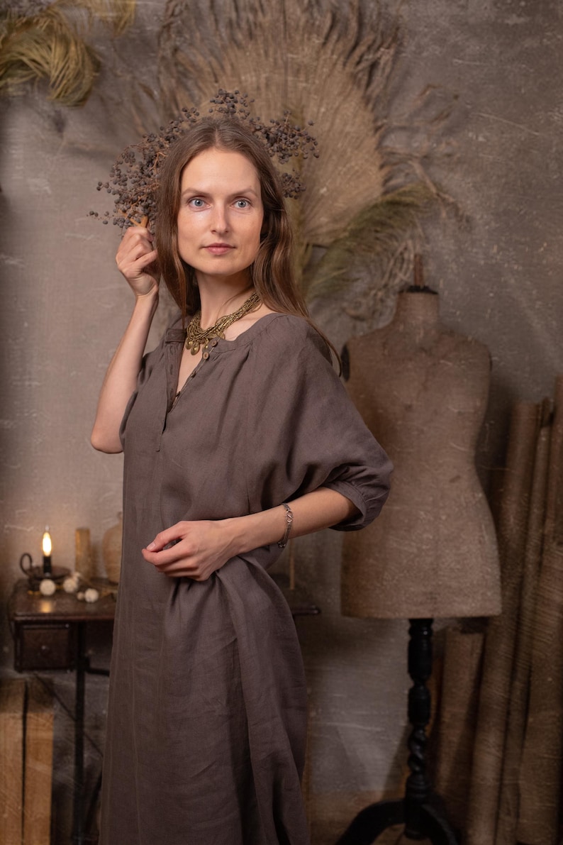 Dark taupe linen dress worn on a model, front three-quarter crop, with round gathered neckline, short button placket, elbow-length puff sleeves, loose body, and visible center front seam.