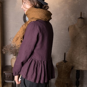 One loose dark plum linen peplum top with long sleeves and a softly gathered waist, shown on a model standing sideways in a knockknocklinen studio.