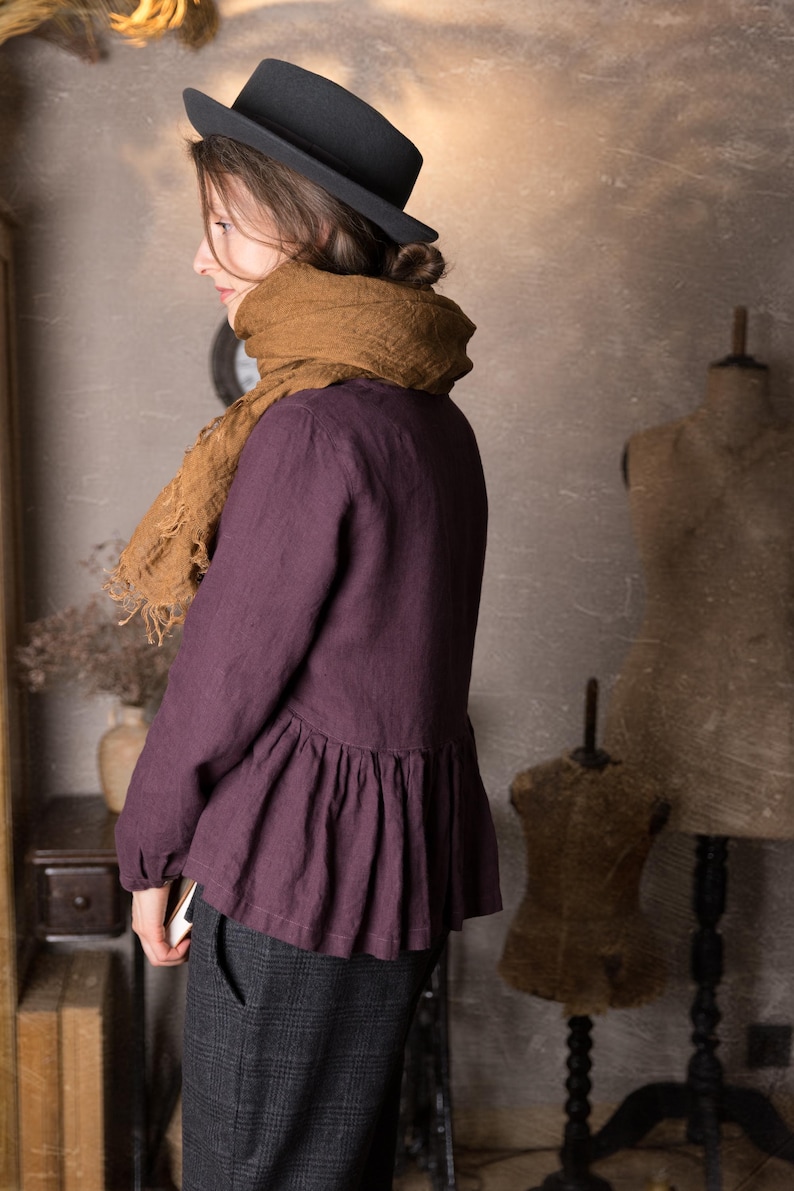 One loose dark plum linen peplum top with long sleeves and a softly gathered waist, shown on a model standing sideways in a knockknocklinen studio.