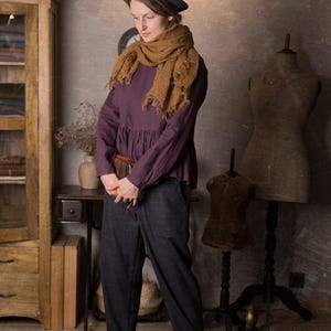 Full-length view of a woman indoors wearing a dark plum long sleeve linen peplum blouse with a gathered hem, styled with grey trousers, brown shoes, a large brown scarf and a black hat.