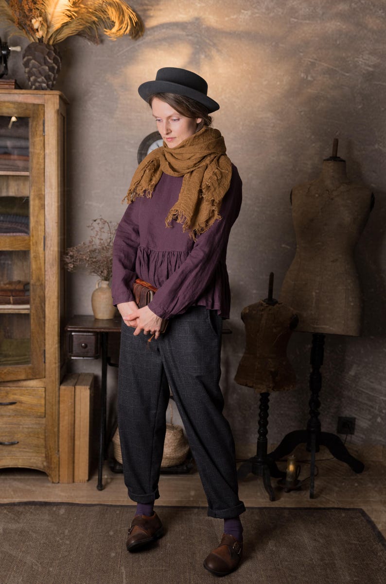 Full-length view of a woman indoors wearing a dark plum long sleeve linen peplum blouse with a gathered hem, styled with grey trousers, brown shoes, a large brown scarf and a black hat.