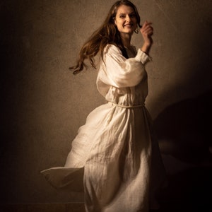 A young woman in a flowing white linen dress with puffed sleeves dances joyfully. Her hair is loose and flowing, and her expression is full of happiness.