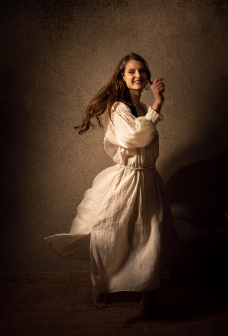 A young woman in a flowing white linen dress with puffed sleeves dances joyfully. Her hair is loose and flowing, and her expression is full of happiness.