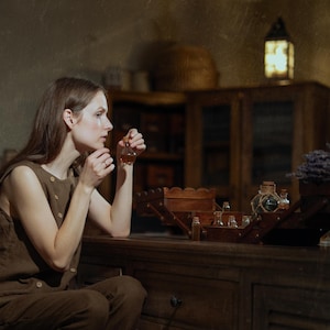 SPARROW linen vest in brown, displayed on a seated model in a side-profile mid-length view, with a sleeveless shape, round neckline, visible light buttons, and a plain side panel, beside wooden trays, small glass bottles, and dried lavender, Knock Knock Linen.