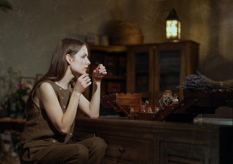 SPARROW linen vest in brown, displayed on a seated model in a side-profile mid-length view, with a sleeveless shape, round neckline, visible light buttons, and a plain side panel, beside wooden trays, small glass bottles, and dried lavender, Knock Knock Linen.