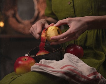 May include: A person peeling a red apple with their hands. The apple is partially peeled and the peel is curled up. There are two other red apples and a brown bowl on a white and red striped cloth.