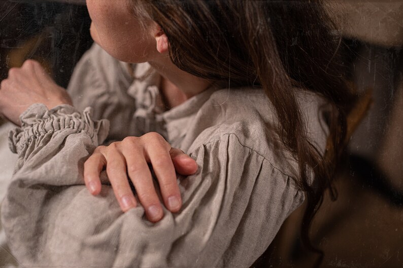 Natural flax linen dress worn by a model, side detail crop, puff sleeve with small pleats at the shoulder seam, wrinkled linen texture, ruffled cuff at wrist, fingers gripping the sleeve, side jaw and ear visible, brown hair draped forward.