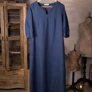 Full-length front view of a deep blue linen dress on a decorative wooden hanger, V-notched neck, elbow-length sleeves, smooth unbroken front panel, lightly wrinkled fabric, hanging from a rustic branch; wooden cabinet at left, knockknocklinen.