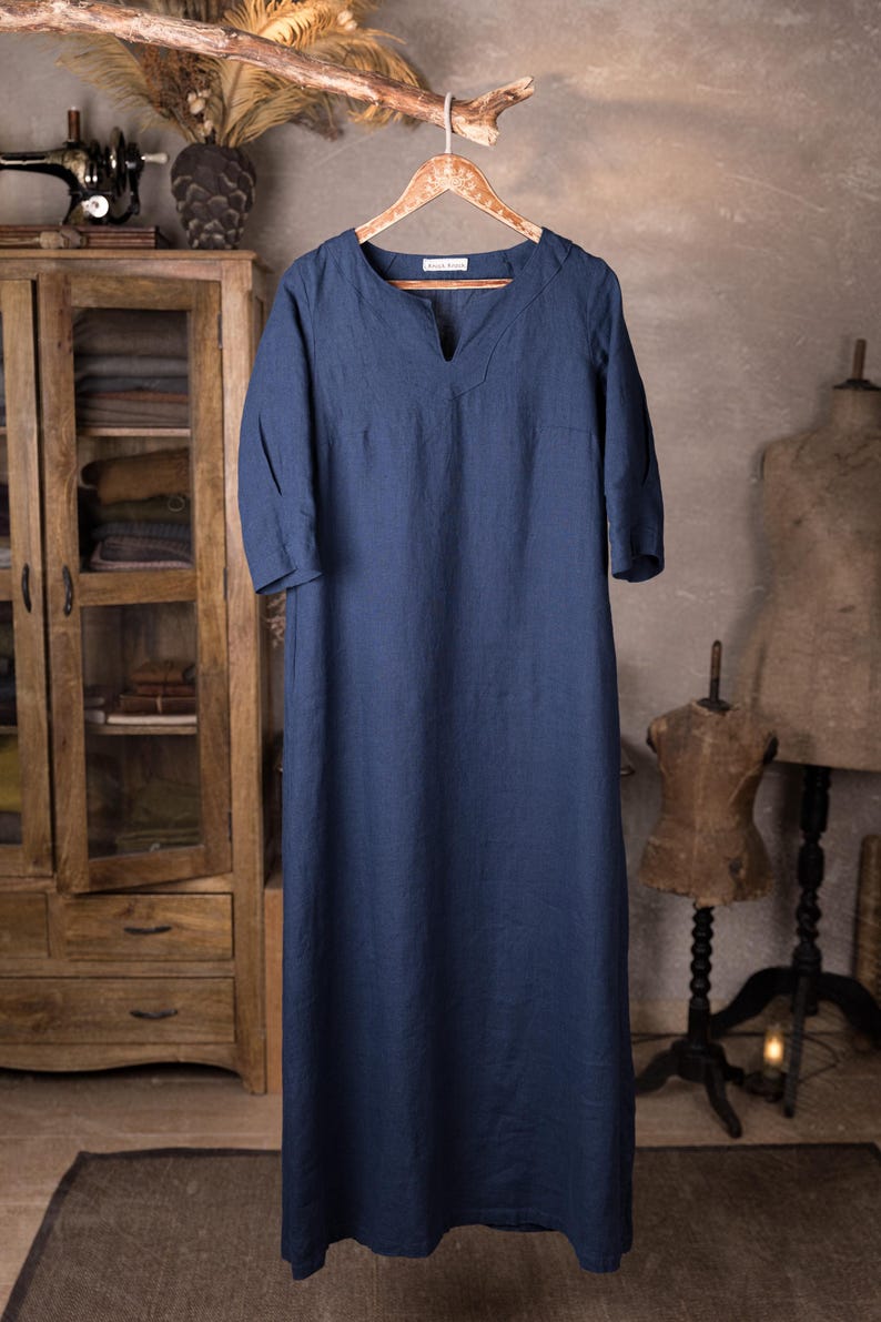 Full-length front view of a deep blue linen dress on a decorative wooden hanger, V-notched neck, elbow-length sleeves, smooth unbroken front panel, lightly wrinkled fabric, hanging from a rustic branch; wooden cabinet at left, knockknocklinen.