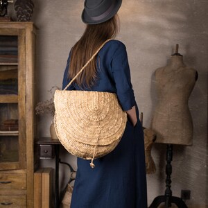 Woman shown from behind in a long dark blue linen dress with sleeves and loose fit, carrying a big straw shoulder bag, standing in a vintage-style room with wooden furniture and dress forms.