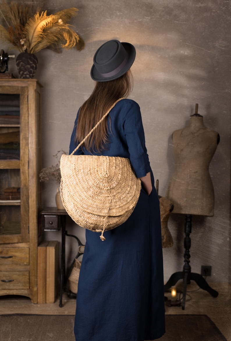 Woman shown from behind in a long dark blue linen dress with sleeves and loose fit, carrying a big straw shoulder bag, standing in a vintage-style room with wooden furniture and dress forms.