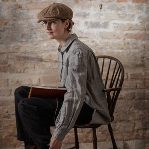 Gray linen shirt with fine dark stripes displayed on a seated model in a full-length three-quarter view, featuring a folded collar, shoulder gathers, long sleeves, broad cuffs, loose body, and rounded hem.