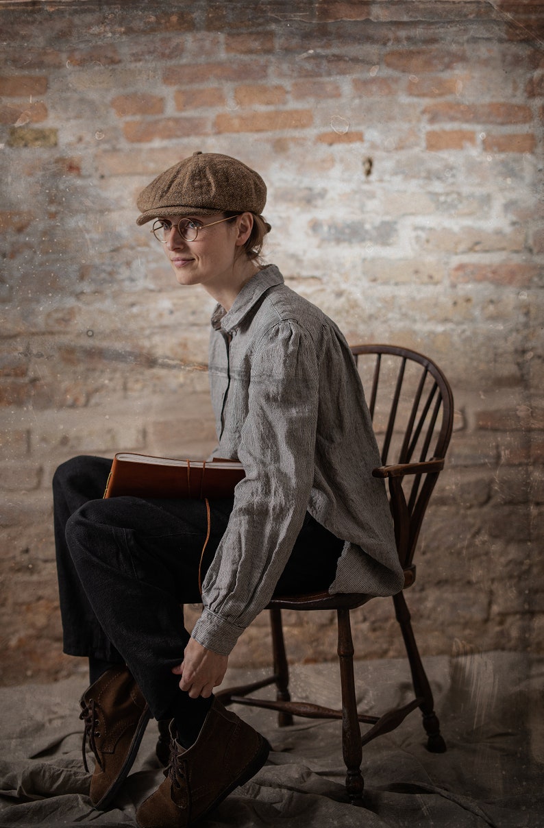 Gray linen shirt with fine dark stripes displayed on a seated model in a full-length three-quarter view, featuring a folded collar, shoulder gathers, long sleeves, broad cuffs, loose body, and rounded hem.