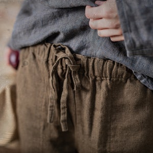 Linen pants in dark brown, shown on a model in a close-up front detail view, with a gathered elastic waistband, loose drawstring ties, visible linen texture, softly wrinkled fabric, and a blue-gray top held up by one hand, Knock Knock Linen.