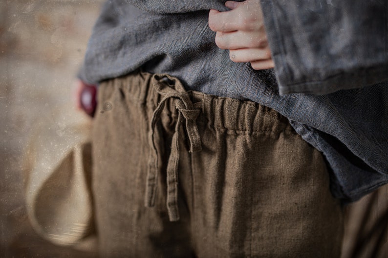 Linen pants in dark brown, shown on a model in a close-up front detail view, with a gathered elastic waistband, loose drawstring ties, visible linen texture, softly wrinkled fabric, and a blue-gray top held up by one hand, Knock Knock Linen.