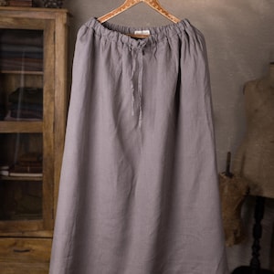 Gray linen skirt shown full-length on a hanger, front view, elasticated gathered waist with drawstring channel, dangling ties, wide A-line shape, plain front with no visible pockets, wooden cabinet and mannequins in background.