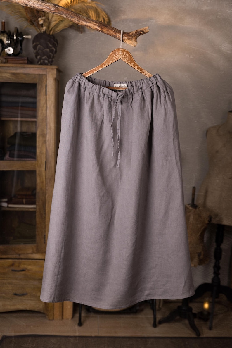 Gray linen skirt shown full-length on a hanger, front view, elasticated gathered waist with drawstring channel, dangling ties, wide A-line shape, plain front with no visible pockets, wooden cabinet and mannequins in background.