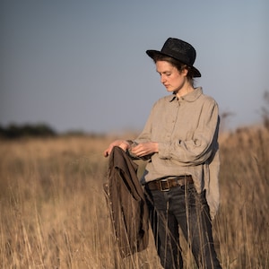 Natural beige linen blouse shown on a standing model in a full-length front view, with a rounded collar, buttoned front, gathered shoulders, long voluminous sleeves, wide cuffs, and a loose hem.