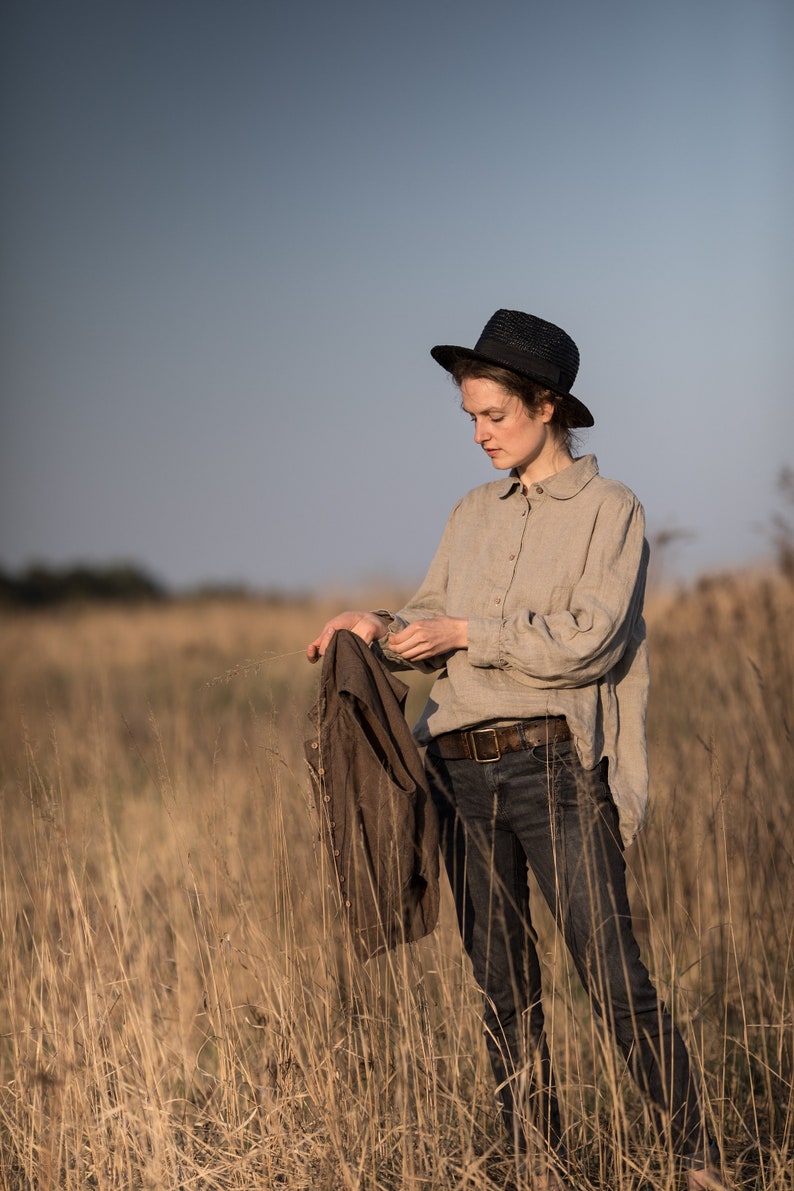 Natural beige linen blouse shown on a standing model in a full-length front view, with a rounded collar, buttoned front, gathered shoulders, long voluminous sleeves, wide cuffs, and a loose hem.