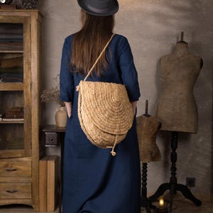 Back view of one woman wearing a dark navy maxi linen dress with 3/4 sleeves and loose kaftan fit, standing indoors with a large woven straw crossbody bag and brown ankle boots.