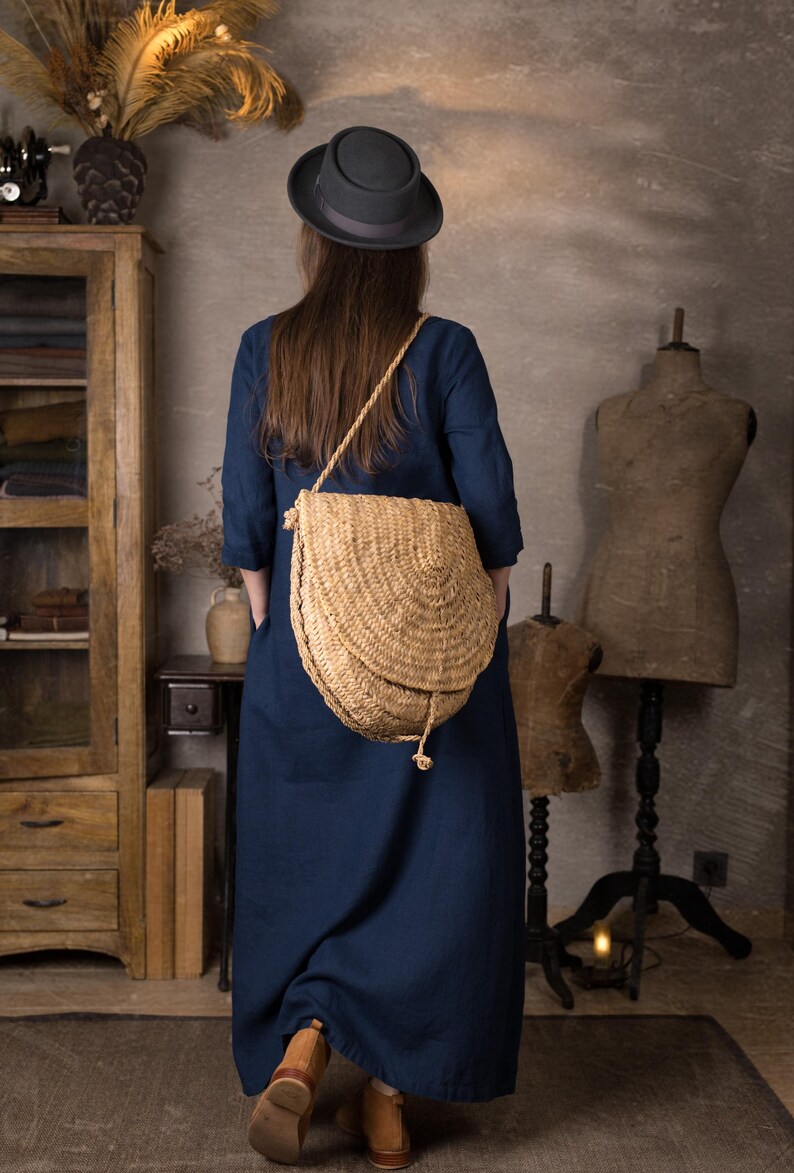 Back view of one woman wearing a dark navy maxi linen dress with 3/4 sleeves and loose kaftan fit, standing indoors with a large woven straw crossbody bag and brown ankle boots.