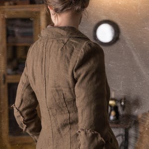 Back view of woman wearing mushroom brown slim fit linen jacket with curved collar, fitted seams and raw elbow patches, shown on model in rustic studio, retro linen jacket by knock knock linen.