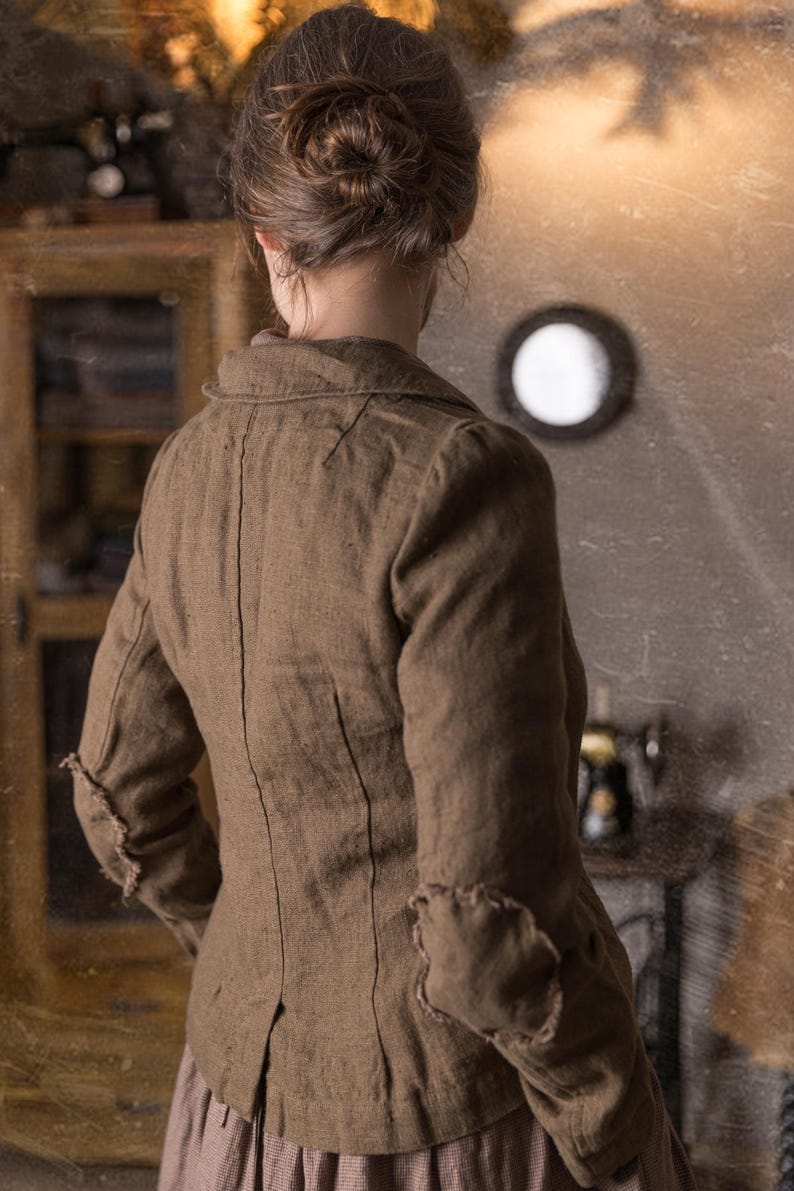Back view of woman wearing mushroom brown slim fit linen jacket with curved collar, fitted seams and raw elbow patches, shown on model in rustic studio, retro linen jacket by knock knock linen.