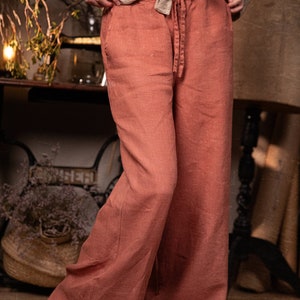 Dusty clay linen wide-leg pants on model, waist-to-feet front view; drawstring tie at center waist, smooth waistband, long wide legs with vertical seams, light wrinkles, hems grazing floor, barefoot.