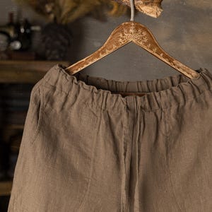 Linen pants in warm mushroom brown colour, displayed on a wooden hanger in a close-up front detail view, with a gathered elastic waistband, center front seam, visible linen texture, softly wrinkled fabric, and a branch above, Knock Knock Linen.