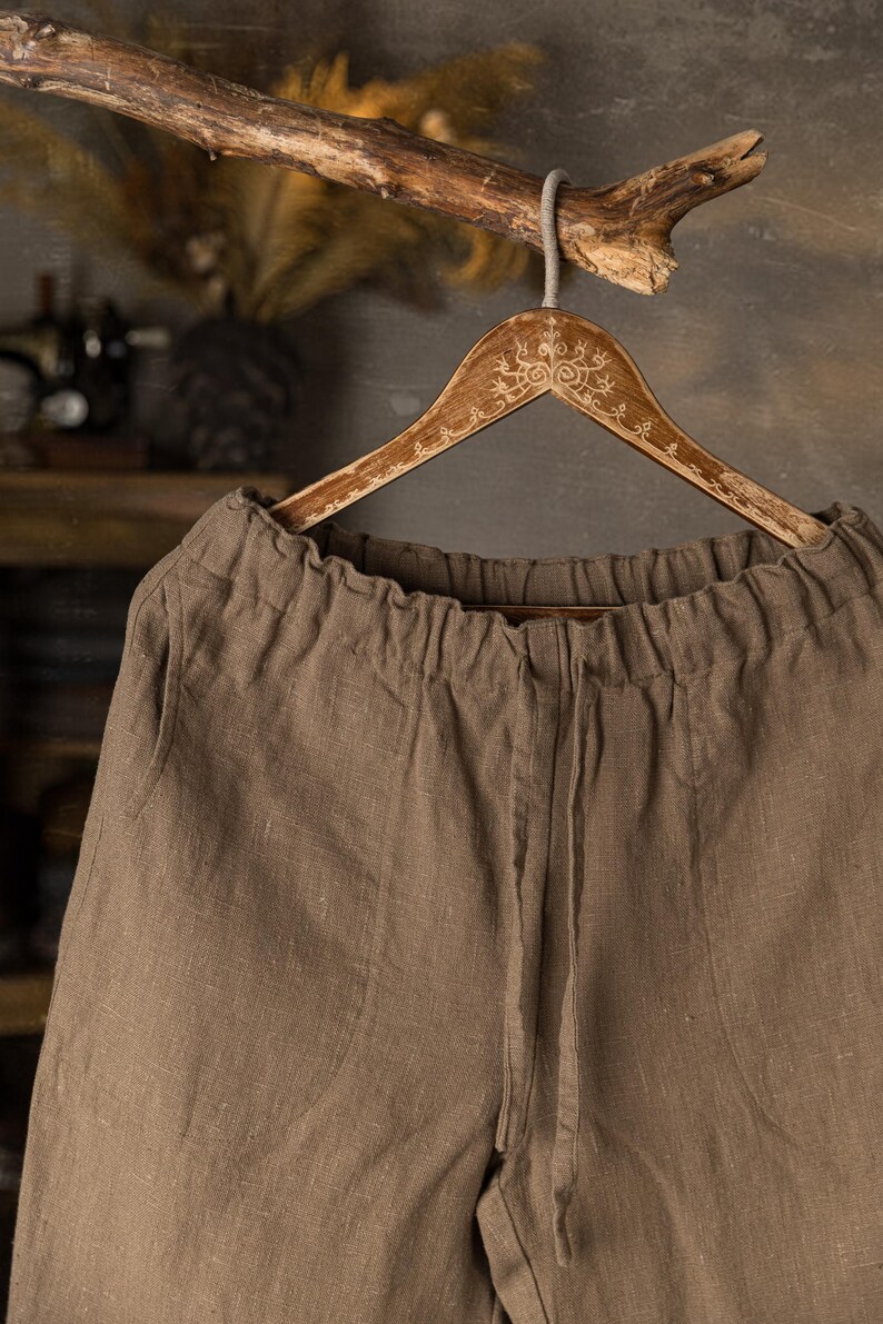 Linen pants in warm mushroom brown colour, displayed on a wooden hanger in a close-up front detail view, with a gathered elastic waistband, center front seam, visible linen texture, softly wrinkled fabric, and a branch above, Knock Knock Linen.