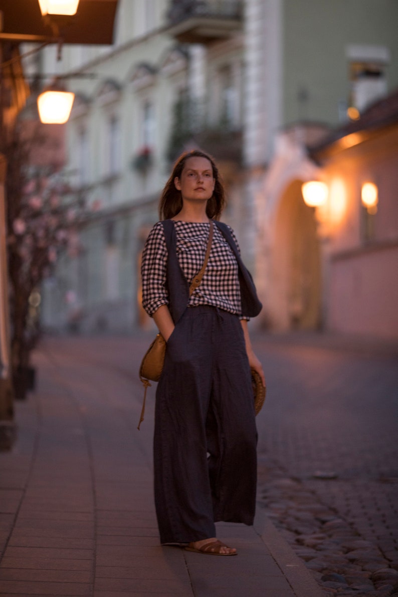 Charcoal linen wide-leg trousers worn on model, full-length three-quarter view; high-rise waistline, wide flowing legs, soft vertical folds, side pocket with hand inside, hem near the ground; evening city sidewalk.