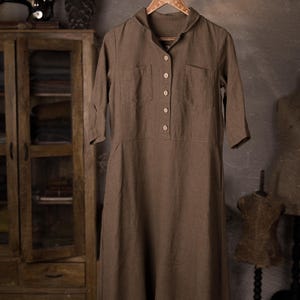 Brown linen dress hanging from a rustic branch on a decorative hanger, front view full-length crop, V-neck collar, short button placket with five buttons, two chest patch pockets, elbow-length sleeves, straight hem, knockknocklinen.