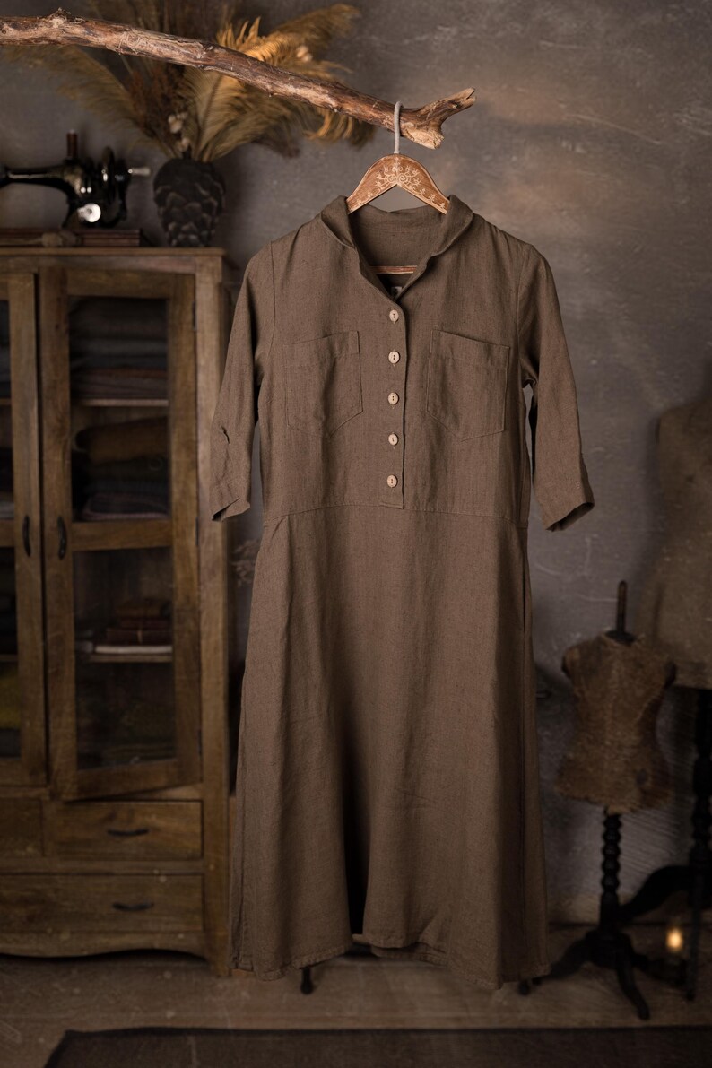Brown linen dress hanging from a rustic branch on a decorative hanger, front view full-length crop, V-neck collar, short button placket with five buttons, two chest patch pockets, elbow-length sleeves, straight hem, knockknocklinen.