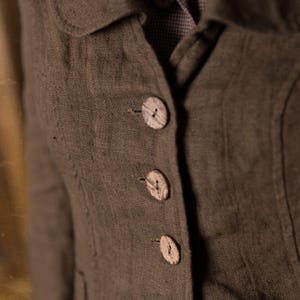 Close-up of mushroom brown slim fit linen jacket on model, showing curved collar, fitted seams and four coconut buttons over checked linen dress, retro buttoned linen jacket by knock knock linen.