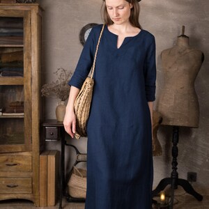 One maxi linen dress in deep navy shown on a woman indoors, ankle length with simple straight silhouette and 3/4 sleeves, styled with a round dark hat and woven shoulder bag against a neutral studio background.