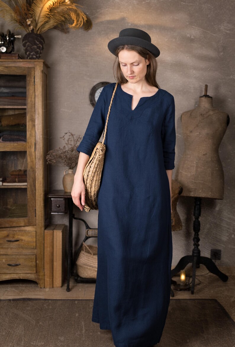 One maxi linen dress in deep navy shown on a woman indoors, ankle length with simple straight silhouette and 3/4 sleeves, styled with a round dark hat and woven shoulder bag against a neutral studio background.